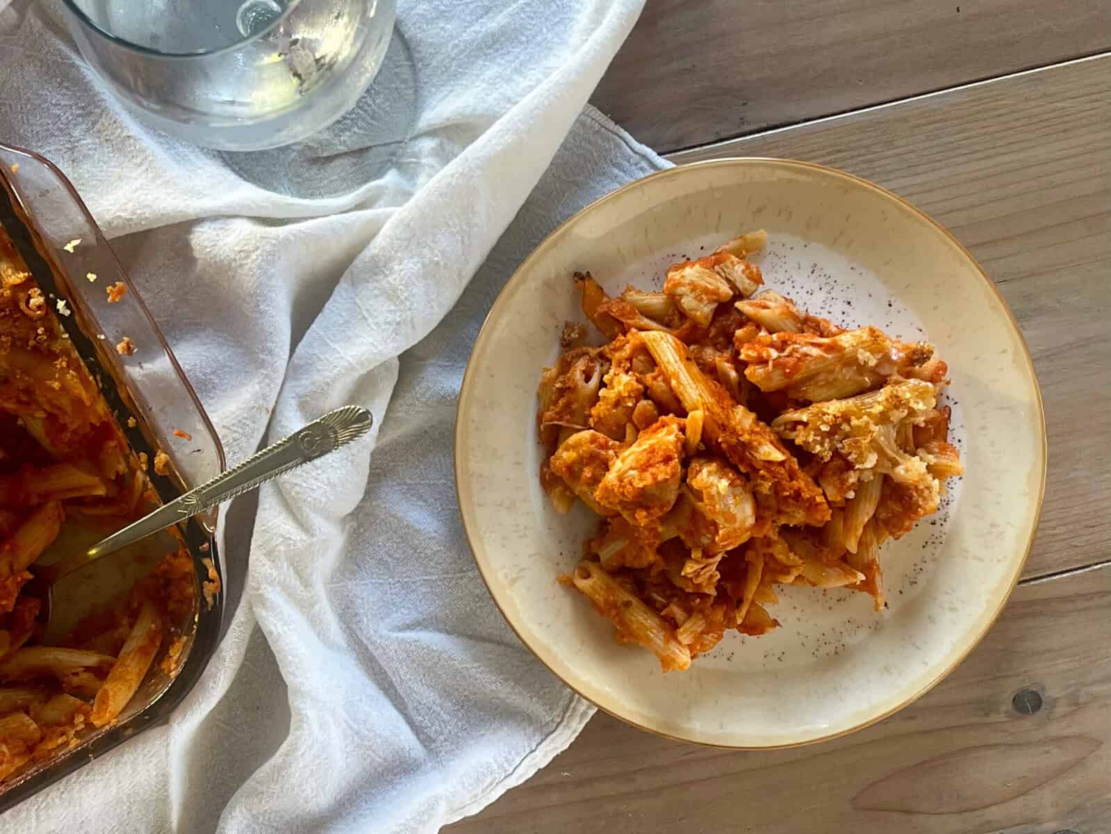 A plate of chicken pasta in tomato sauce with breadcrumb streusel next to a water glass and casserole